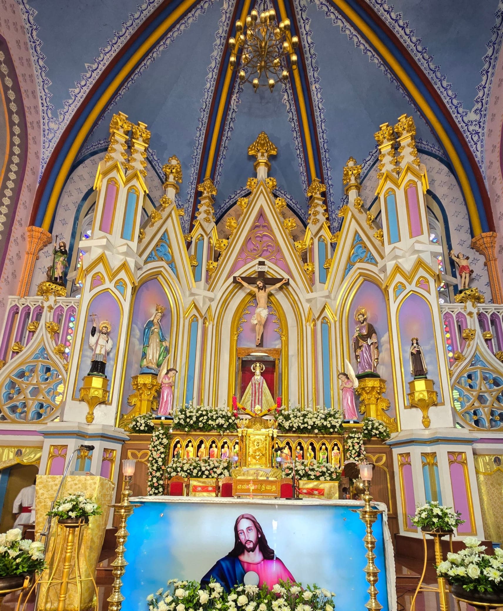 The high altar: a five-bay carved screen rising to gilded spires under the painted vault, the crucifix on its centre axis, and twelve small statues in a gold arcade either side of the tabernacle