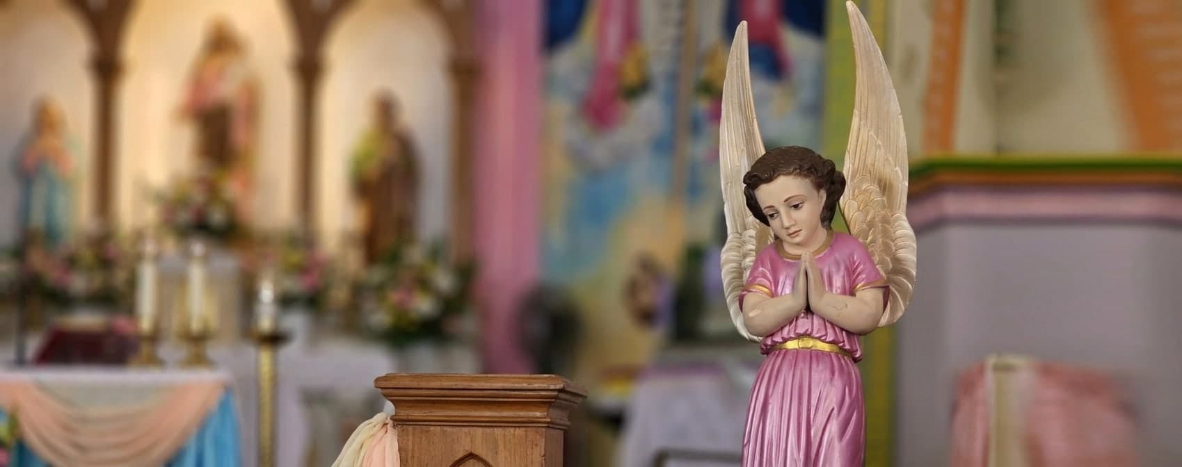 A carved angel in a rose-coloured robe kneels with hands joined in prayer beside the pulpit of the shrine, the flower-dressed high altar and its saints out of focus behind