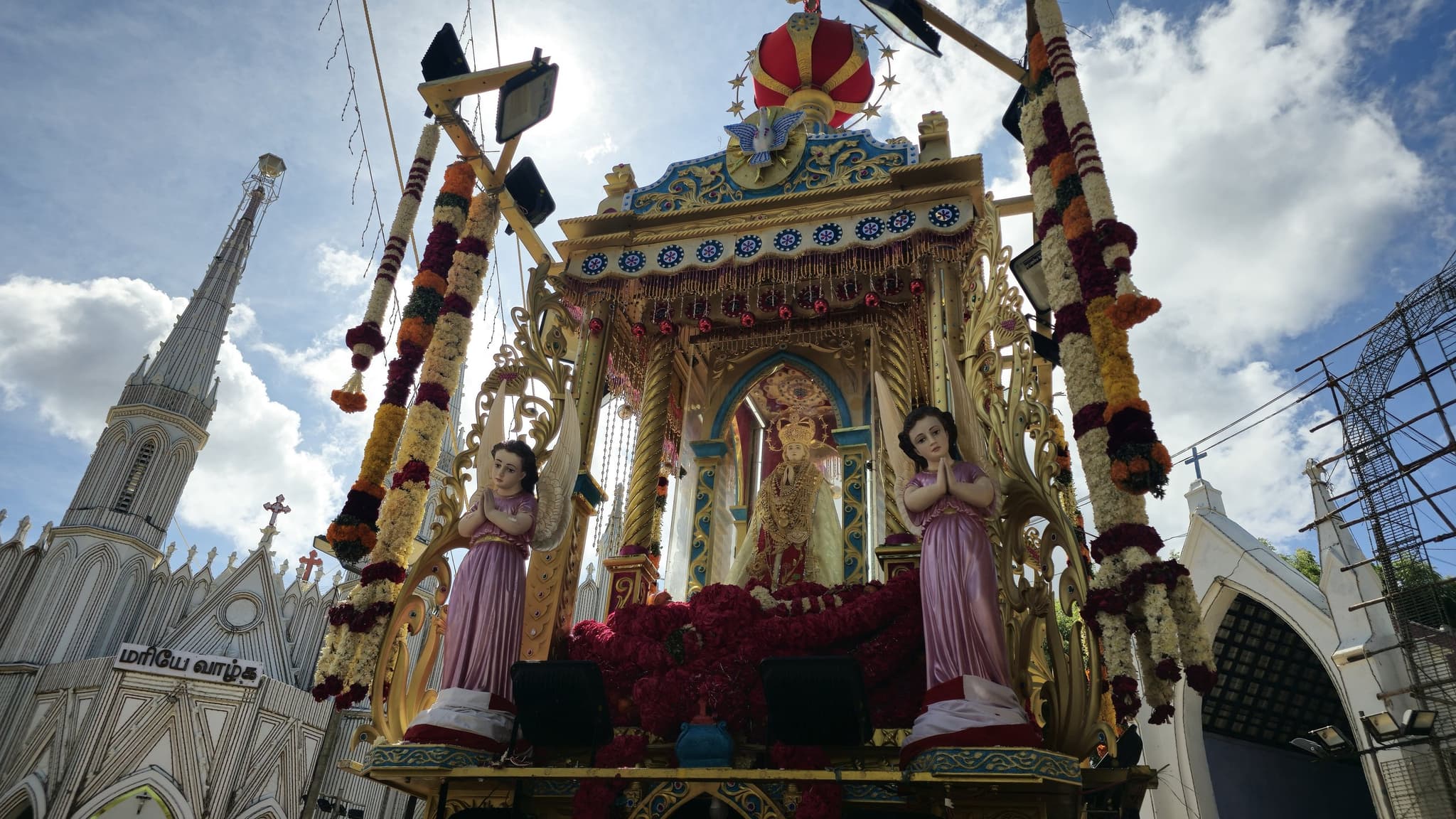 The crowned statue of Our Lady of the Assumption on her flower-decked processional chariot during the August feast, an angel kneeling either side and the church spire rising behind